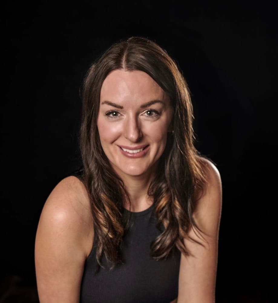 Smiling woman with long brown hair wearing a black sleeveless top against a black background.